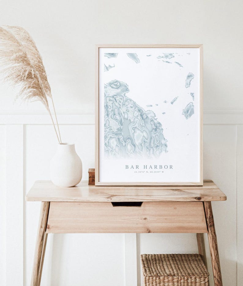 May include: A framed print of a blue and white map of Bar Harbor, Maine. The map is in a light wood frame and is sitting on a wooden table.