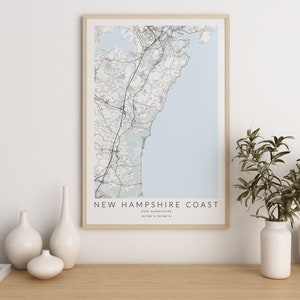 K&ouml;nnte beinhalten: Eine gerahmte Schwarzwei&szlig;karte der K&uuml;ste von New Hampshire mit dem Text "NEW HAMPSHIRE COAST" und "NEW HAMPSHIRE" in kleinerer Schrift darunter. Die Karte hat einen hellblauen Hintergrund und ist von einem hellen Holzrahmen umgeben. Die Karte liegt auf einem hellen Holztisch mit drei wei&szlig;en Keramikvasen und einem Buch.