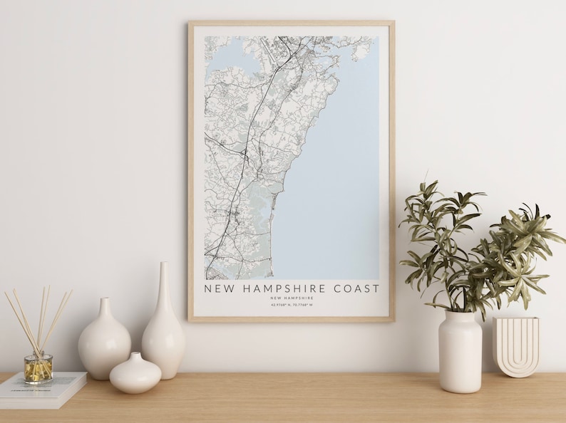 K&ouml;nnte beinhalten: Eine gerahmte Schwarzwei&szlig;karte der K&uuml;ste von New Hampshire mit dem Text "NEW HAMPSHIRE COAST" und "NEW HAMPSHIRE" in kleinerer Schrift darunter. Die Karte hat einen hellblauen Hintergrund und ist von einem hellen Holzrahmen umgeben. Die Karte liegt auf einem hellen Holztisch mit drei wei&szlig;en Keramikvasen und einem Buch.
