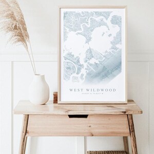 May include: A framed map print of West Wildwood in light blue and white tones. The print is displayed on a wooden table with a white vase and decorative dried grass. The table has a small drawer and a woven basket underneath.
