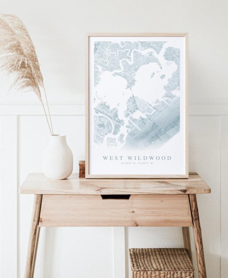 May include: A framed map print of West Wildwood in light blue and white tones. The print is displayed on a wooden table with a white vase and decorative dried grass. The table has a small drawer and a woven basket underneath.