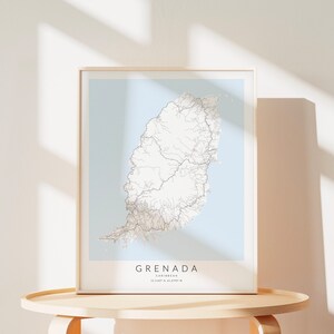 May include: A framed map of Grenada, a Caribbean island, with a light blue background and white lines representing roads. The frame is light wood and the map is on a light wood table.