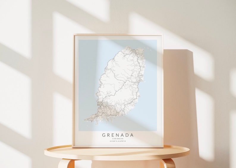 May include: A framed map of Grenada, a Caribbean island, with a light blue background and white lines representing roads. The frame is light wood and the map is on a light wood table.