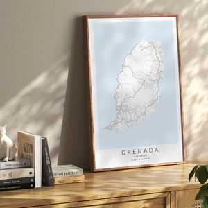 May include: A framed map of Grenada, a Caribbean island, with a light blue background and white lines representing roads. The map is on a wooden table with a white ceramic cat figurine, a stack of books, and a vase with a green plant.