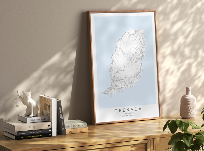 May include: A framed map of Grenada, a Caribbean island, with a light blue background and white lines representing roads. The map is on a wooden table with a white ceramic cat figurine, a stack of books, and a vase with a green plant.