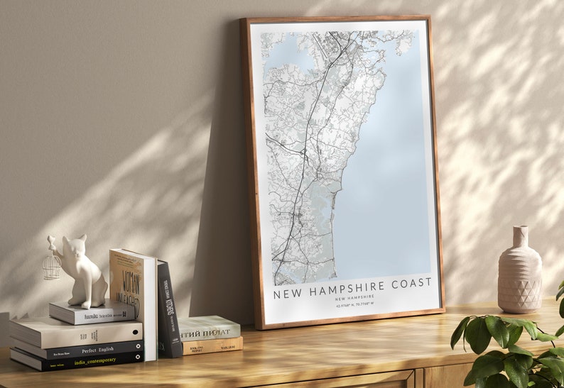 K&ouml;nnte beinhalten: Ein gerahmter Kartenabdruck der K&uuml;ste von New Hampshire mit hellblauem Hintergrund und schwarzen Linien, die die Stra&szlig;en und die K&uuml;ste zeigen. Der Text "NEW HAMPSHIRE COAST" ist in Schwarz am unteren Rand der Karte gedruckt.