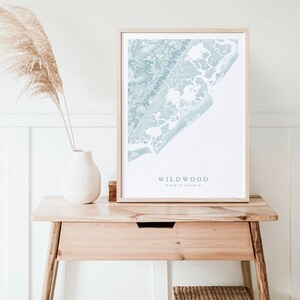Może przedstawiać: Wydrukowana mapa Wildwood w stanie New Jersey, w niebieskich i białych kolorach, oprawiona w jasną drewnianą ramę. Mapa stoi na drewnianym stole, obok białego wazonu i wiklinowego koszyka.