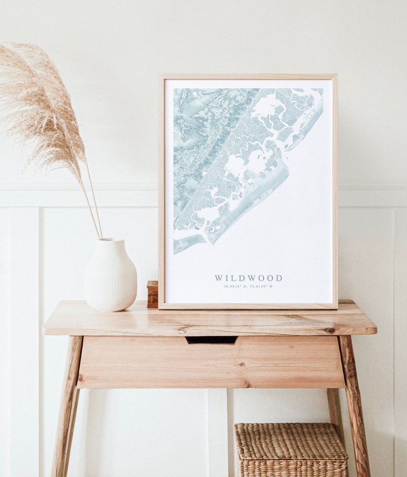 Może przedstawiać: Wydrukowana mapa Wildwood w stanie New Jersey, w niebieskich i białych kolorach, oprawiona w jasną drewnianą ramę. Mapa stoi na drewnianym stole, obok białego wazonu i wiklinowego koszyka.