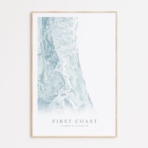 May include: A framed print featuring a coastal map in shades of blue and white. The artwork is labeled "First Coast" with coordinates below. The print has a light wood frame and is displayed against a white wall.