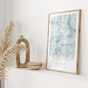 May include: A framed print of a map of London in shades of blue and green. The map is surrounded by a light wood frame. The print is on a white shelf with a wooden vase and bowls.