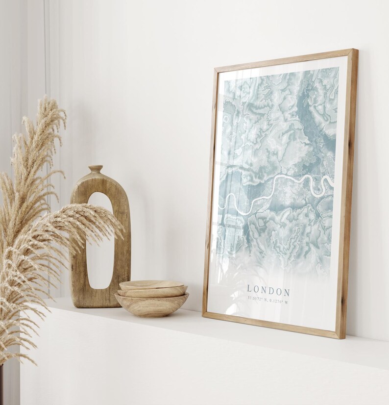 May include: A framed print of a map of London in shades of blue and green. The map is surrounded by a light wood frame. The print is on a white shelf with a wooden vase and bowls.
