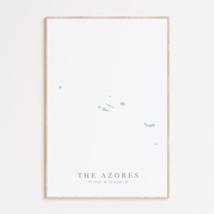 May include: A minimalist print of the Azores islands in a light blue color on a white background. The print is framed in a light wood frame. The text "THE AZORES" and the coordinates "37.7413° N, 25.6764° W" are printed below the map.