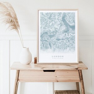 May include: A framed print of a blue and white map of London, England. The map shows the city's streets and waterways. The text "LONDON" and the coordinates "51.5074&deg; N, 0.1278&deg; W" are printed below the map.