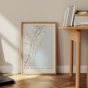 K&ouml;nnte beinhalten: Ein gerahmter Kartenabdruck der K&uuml;ste von New Hampshire mit hellblauem Hintergrund und schwarzen Linien, die die Stra&szlig;en und die K&uuml;ste zeigen. Der Rahmen ist in hellem Holz gehalten. Der Text auf der Karte lautet "New Hampshire Coast, New Hampshire, 43.09&deg;N, 70.71&deg;W".