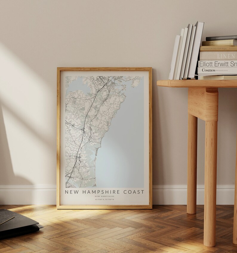 K&ouml;nnte beinhalten: Ein gerahmter Kartenabdruck der K&uuml;ste von New Hampshire mit hellblauem Hintergrund und schwarzen Linien, die die Stra&szlig;en und die K&uuml;ste zeigen. Der Rahmen ist in hellem Holz gehalten. Der Text auf der Karte lautet "New Hampshire Coast, New Hampshire, 43.09&deg;N, 70.71&deg;W".