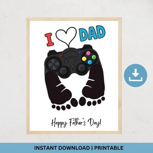 May include: A framed print with a white background features a black video game controller design with the words "I ❤️ DAD" in red and blue. Below the controller are two black footprints, and the words "Happy Father's Day!" are written in black.