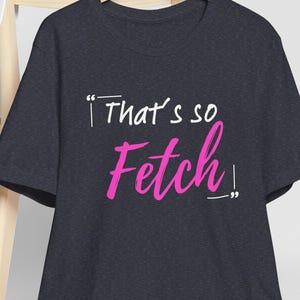 Może przedstawiać: Ciemnoszara koszulka z napisem "That's so Fetch!" w białych i różowych literach. Biała czapka z daszkiem spoczywa na drewnianym krześle w tle. Koszulka wisi na drewnianym wieszaku.
