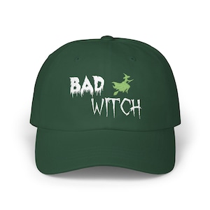Op de afbeelding: Een donkergroene baseballcap met de woorden "BAD WITCH" in witte, verweerde letters. Een groene silhouet van een heks op een bezem staat boven het woord "WITCH". De cap heeft een gebogen klep en een gestructureerde kroon.