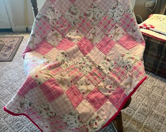 Puede incluir: Una colcha de retazos rosa y blanca con estampado de ciervo y flores. La colcha está doblada y drapeada sobre una mecedora.