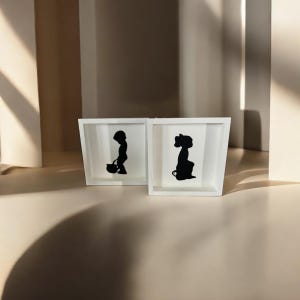 Könnte beinhalten: Zwei weiß gerahmte Kunstwerke mit schwarzen Silhouetten. Eines zeigt eine Person, die in einen Eimer uriniert, das andere zeigt eine Person, die auf einer Toilette sitzt. Die Kunstwerke werden auf einer hellfarbigen Oberfläche ausgestellt.