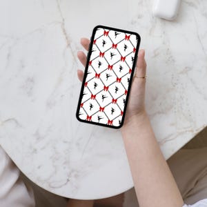 以下が含まれることがあります： スマートフォンには、黒いバレリーナのシルエット、赤いリボン、チェーンリンクが白い背景に繰り返されるパターンが表示されています。 金色の指輪をした手でスマートフォンを持っており、大理石の表面に置かれています。