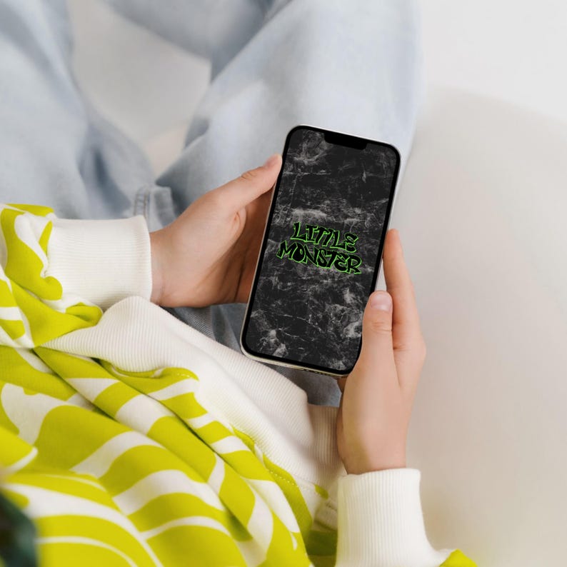 May include: A smartphone displaying a black marble-textured background with the text LITTLE MONSTER in bright green graffiti-style lettering. The device is held in front of a white and lime green patterned sweatshirt and light blue denim fabric.