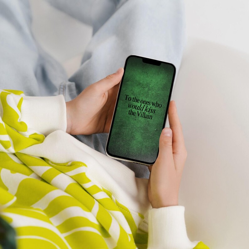 May include: A smartphone displaying a dark green textured background with the black text "To the ones who would kiss the Villain" centered on the screen. The device is held in a neutral position, showcasing the screen's digital graphic design.