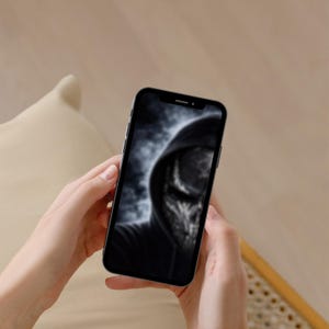以下が含まれることがあります： 黒いフード付きスウェットシャツを着た人物のぼやけた画像を表示するスマートフォン。 携帯電話は、ニュートラルな背景に対して両手で持たれています。 画面にはグレースケール画像が表示されます。