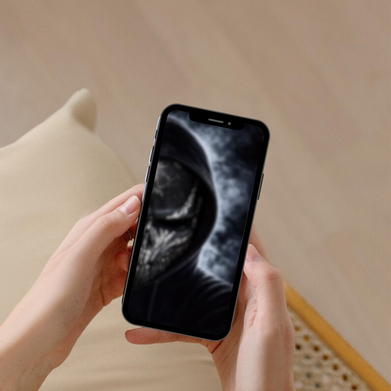 以下が含まれることがあります： 黒いスマートフォン。フード付きの人物のぼやけた画像が表示され、頭蓋骨のような顔をしています。 携帯電話は、ニュートラルな背景に対して両手で持たれています。