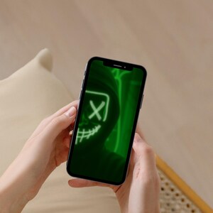 May include: A smartphone displaying a vibrant green digital graphic on its screen. The image features a stylized, glowing neon-style mask with an X-shaped eye and a stitched mouth design, set against a dark, shadowy background within the display.
