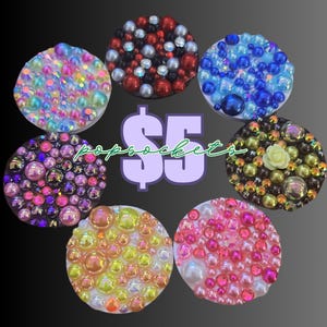 May include: A collection of bedazzled PopSockets in various colors, including pink, blue, and gold. Each round PopSocket is covered in beads and gems. The image also includes the text "$5" and "popsockets".