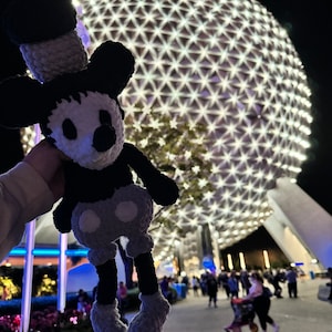 May include: A black and white crocheted Mickey Mouse doll with a grey hat and trousers, held in front of a large, illuminated sphere with a geometric pattern.