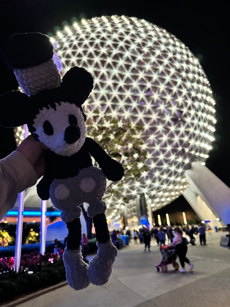 May include: A black and white crocheted Mickey Mouse doll with a grey hat and trousers, held in front of a large, illuminated sphere with a geometric pattern.