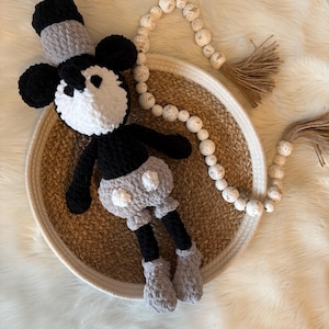 May include: A crocheted black and white Mickey Mouse plush toy with grey accents sits in a woven basket on a white faux fur surface. A white beaded garland with a brown tassel hangs in the background.