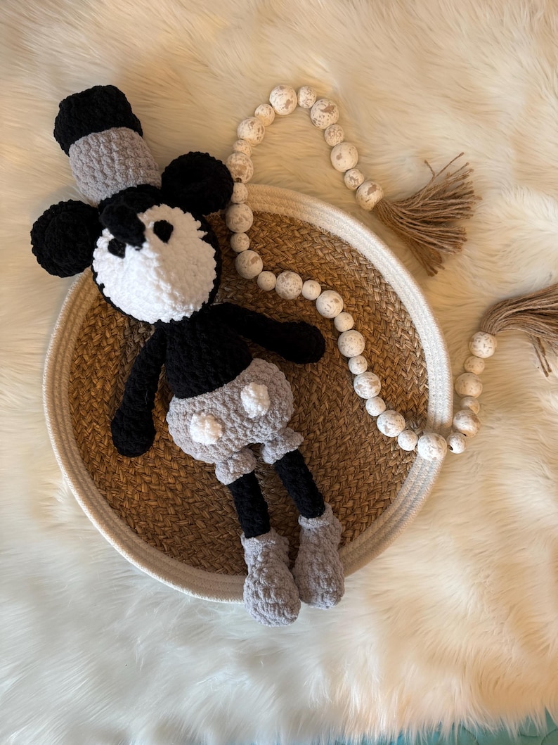 May include: A crocheted black and white Mickey Mouse plush toy with grey accents sits in a woven basket on a white faux fur surface. A white beaded garland with a brown tassel hangs in the background.