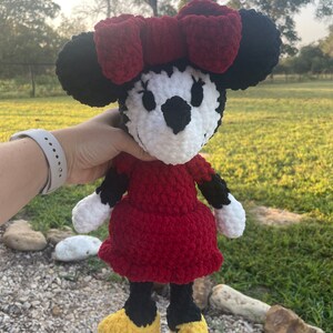 May include: A crocheted plush toy of Minnie Mouse. The toy is red, black, white, and yellow. Minnie Mouse is wearing a red dress and a red bow.