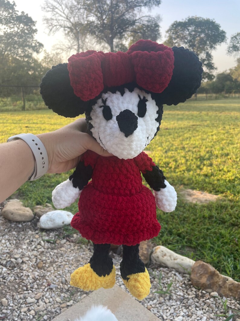 May include: A crocheted plush toy of Minnie Mouse. The toy is red, black, white, and yellow. Minnie Mouse is wearing a red dress and a red bow.