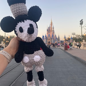 May include: A crocheted Mickey Mouse plush toy in black, white and grey, with a black hat and large eyes. The toy is being held in front of a blurred background of a castle.