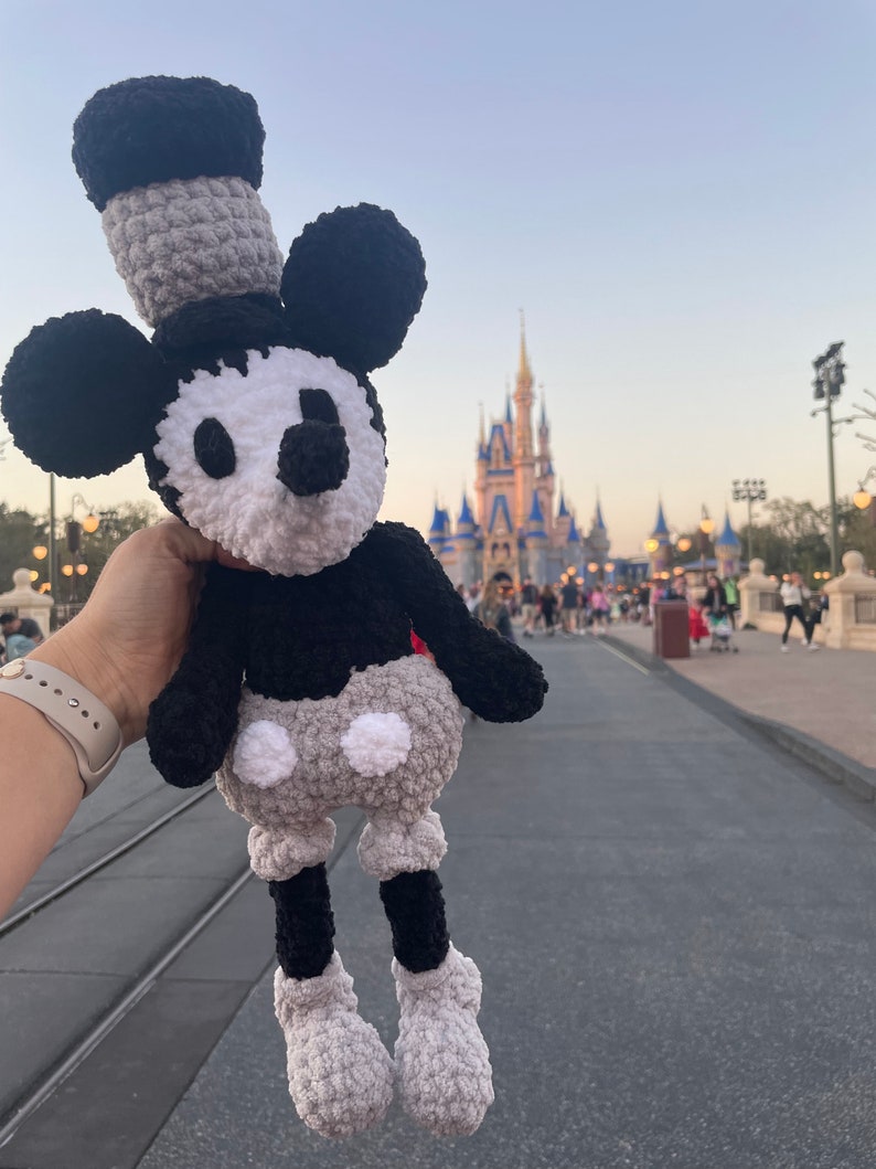May include: A crocheted Mickey Mouse plush toy in black, white and grey, with a black hat and large eyes. The toy is being held in front of a blurred background of a castle.