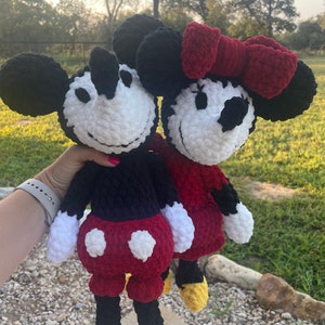 May include: A crocheted Mickey Mouse and Minnie Mouse plush toy set. Mickey is wearing black trousers and red shorts with white polka dots. Minnie is wearing a red dress with a red bow. Both have white faces with black eyes and noses.