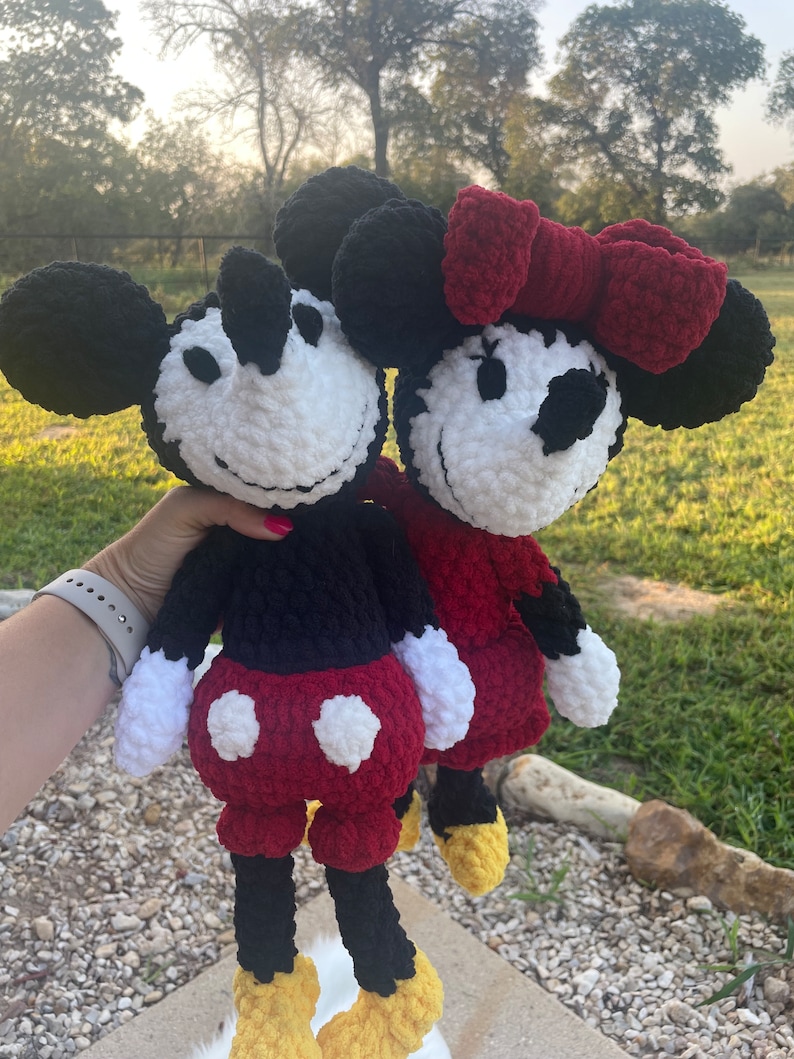 May include: A crocheted Mickey Mouse and Minnie Mouse plush toy set. Mickey is wearing black trousers and red shorts with white polka dots. Minnie is wearing a red dress with a red bow. Both have white faces with black eyes and noses.