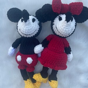 May include: A crocheted Mickey Mouse and Minnie Mouse plush toy set. Mickey is wearing red shorts with white polka dots and yellow shoes. Minnie is wearing a red dress with a red bow and yellow shoes.
