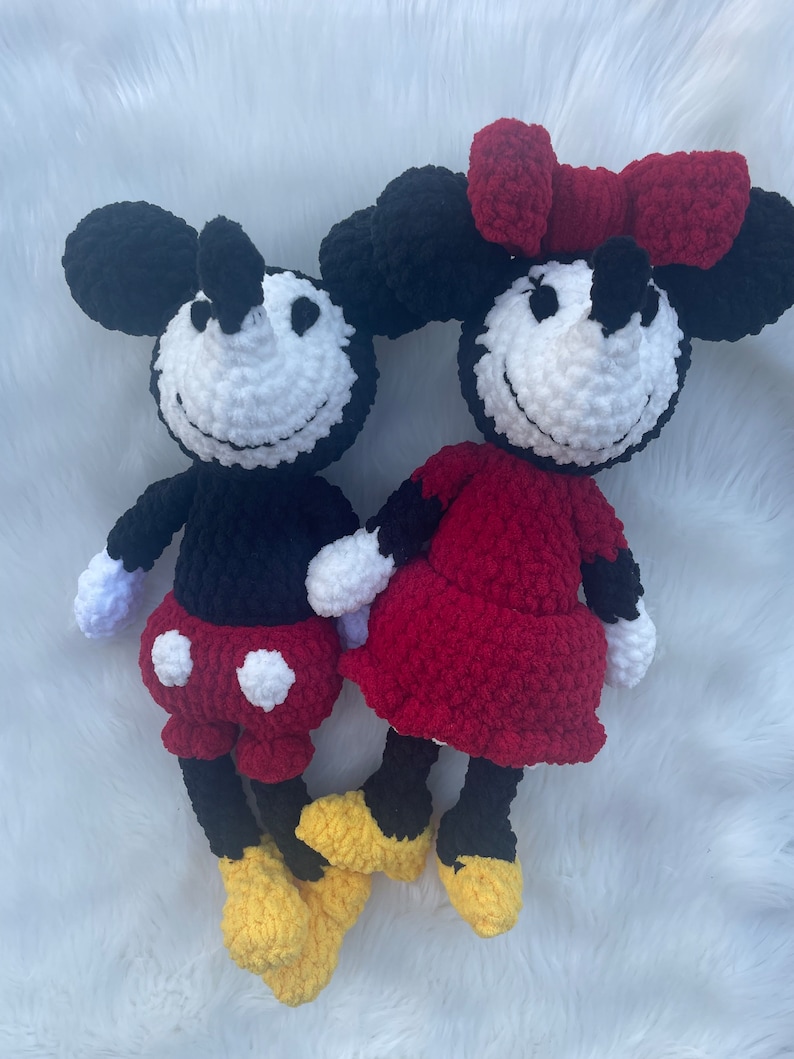 May include: A crocheted Mickey Mouse and Minnie Mouse plush toy set. Mickey is wearing red shorts with white polka dots and yellow shoes. Minnie is wearing a red dress with a red bow and yellow shoes.