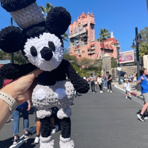 May include: A black and white crocheted Mickey Mouse plush toy with a grey hat. The toy is being held by a hand in front of a pink building with a tower.