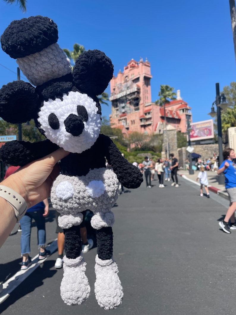 May include: A black and white crocheted Mickey Mouse plush toy with a grey hat. The toy is being held by a hand in front of a pink building with a tower.
