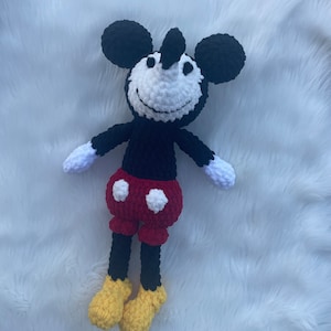 May include: A crocheted plush toy of Mickey Mouse. The toy is black with white eyes and a white smile. It is wearing red shorts with white polka dots and yellow shoes.