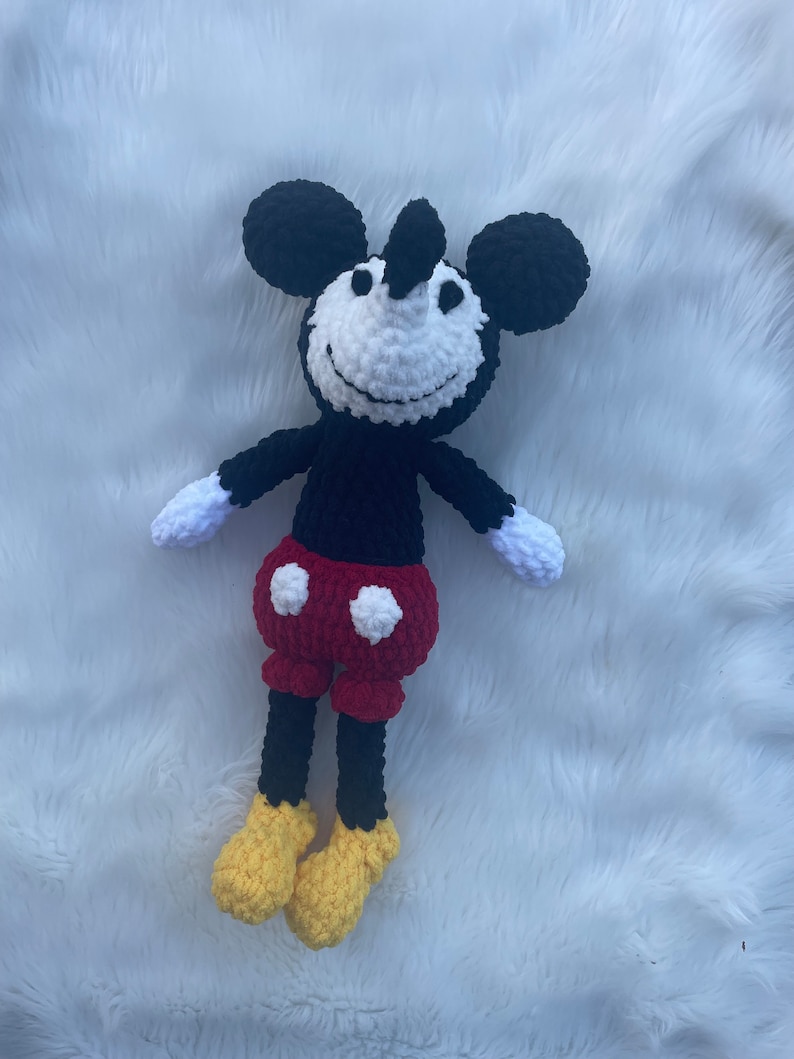 May include: A crocheted plush toy of Mickey Mouse. The toy is black with white eyes and a white smile. It is wearing red shorts with white polka dots and yellow shoes.