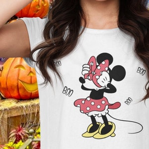 May include: A white short-sleeved t-shirt featuring a graphic of Minnie Mouse covering her eyes with her hands. She wears a red polka-dot dress and matching bow, with yellow shoes. The word BOO is printed in black text around the character.