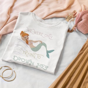 Puede incluir: Camiseta blanca con un gráfico de una sirena con cabello rojo y una corona. El texto "We were only trying to drown her" está impreso en la camiseta.
