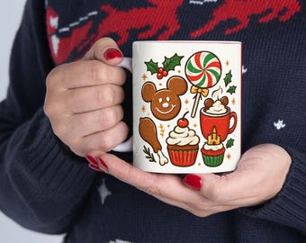 May include: White ceramic mug with a red interior, featuring a festive design of gingerbread, cupcakes, a lollipop, and a mug of hot cocoa. The mug is held by a person wearing a blue sweater.